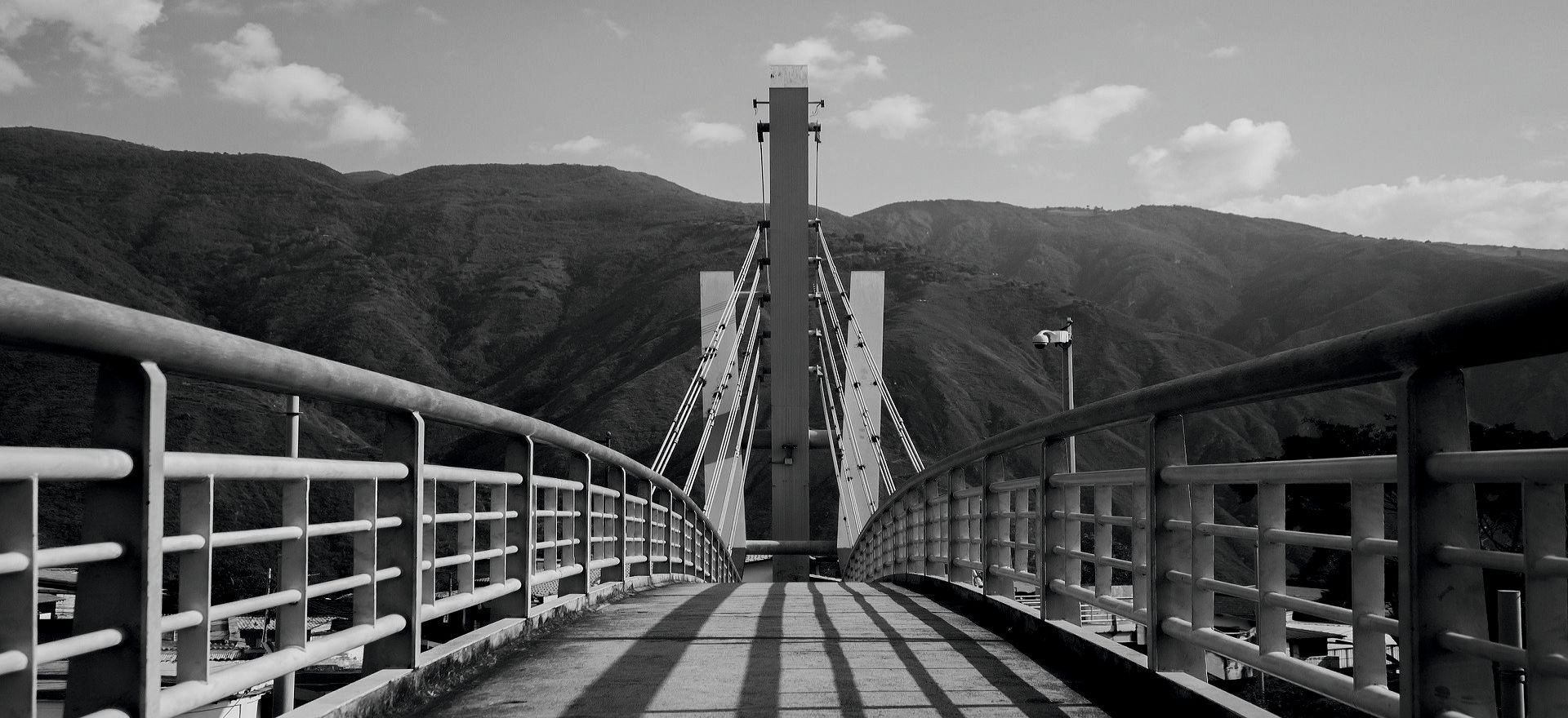 A cable-stayed footbridge leading towards mountains, photographed in black and white.